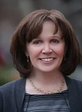 Joan C. Waters is smiling directly into camera wearing a dark sweater with a dark striped boat neck blouse underneath. Also, she is wearing a gold linked necklace.